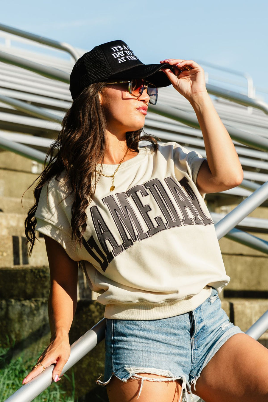 A woman in sunglasses adjusts her black cap, wearing the Panache Apparel Collection's Beige & Gray Game Day Short Sleeve Sweatshirt styled with ripped denim shorts, as she stands by metal bleachers in sunlight.