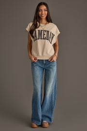 A woman stands smiling against a plain gray background, wearing the Panache Apparel Collection Beige & Gray Game Day Short Sleeve Sweatshirt, blue wide-leg jeans, brown shoes, and gold jewelry.