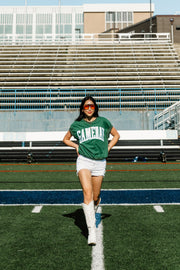 A person wearing the Panache Apparel Collection Green & White Game Day Short Sleeve Sweatshirt, paired with white shorts and white boots, walks confidently on a football field with empty bleachers in the background.