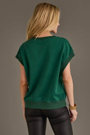 A woman with long blonde hair stands facing away, wearing the Panache Apparel Collection Green & White Game Day Short Sleeve Sweatshirt paired with black leather pants and accessorized with a chunky gold bracelet.