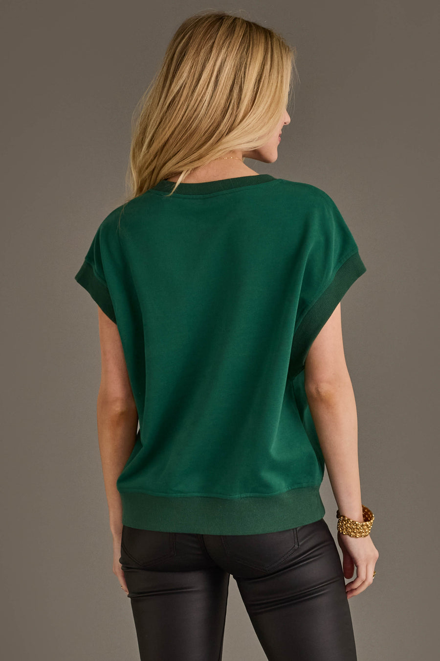 A woman with long blonde hair stands facing away, wearing the Panache Apparel Collection Green & White Game Day Short Sleeve Sweatshirt paired with black leather pants and accessorized with a chunky gold bracelet.