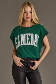 A woman wearing the Panache Apparel Collection's Green & White Game Day Short Sleeve Sweatshirt and black leather pants stands against a plain background, smiling slightly with one hand in her pocket.