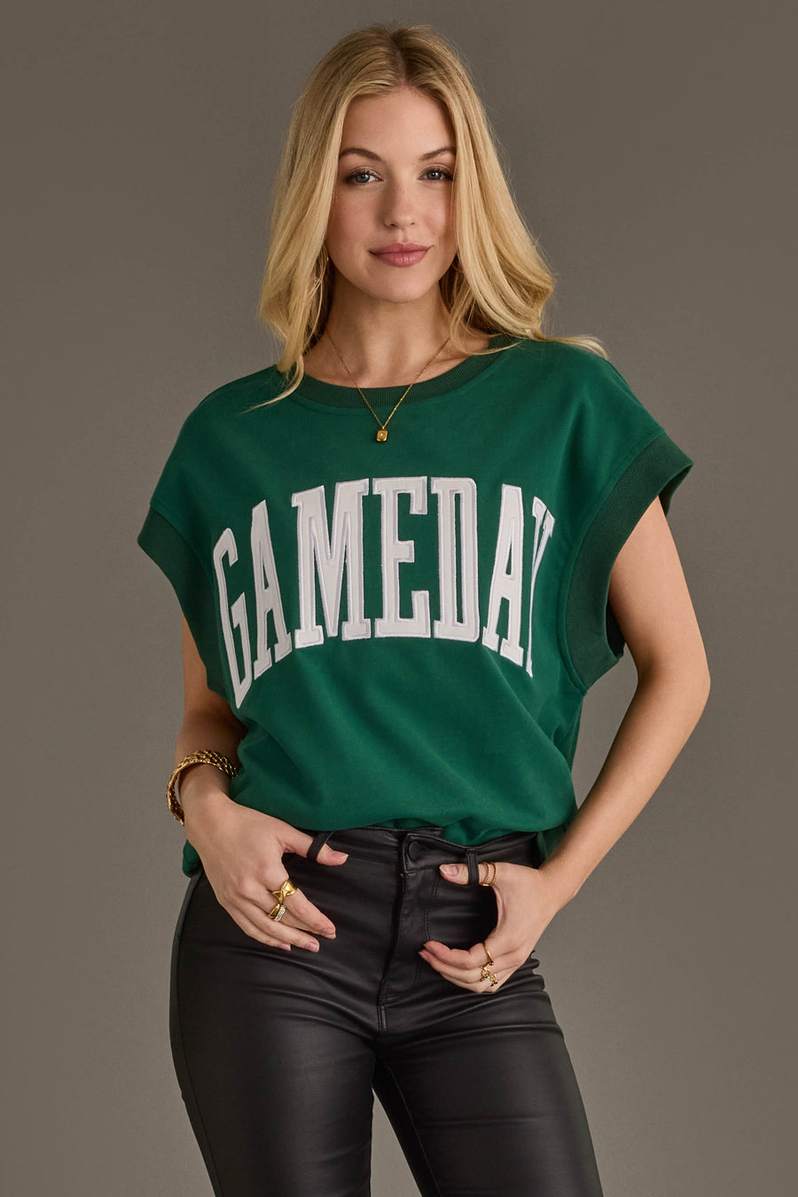 A woman wearing the Panache Apparel Collection's Green & White Game Day Short Sleeve Sweatshirt and black leather pants stands against a plain background, smiling slightly with one hand in her pocket.