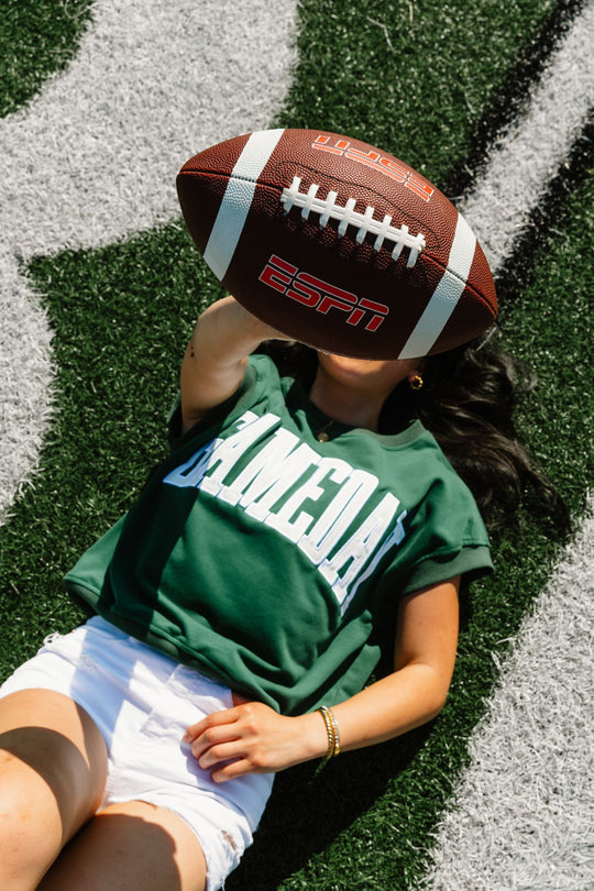 A person lying on a football field holds an ESPN football in front of their face, wearing the Panache Apparel Collection Green & White Game Day Short Sleeve Sweatshirt and white shorts.