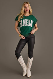A woman poses against a plain backdrop wearing the Panache Apparel Collection Green & White Game Day Short Sleeve Sweatshirt, paired with black pants and white cowboy boots. She has long blonde hair and accessorizes with a bracelet and necklace.