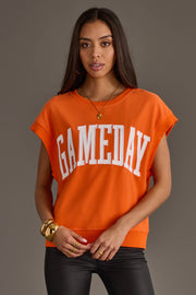 A woman with long dark hair wears the Panache Apparel Collection Orange & White Game Day Short Sleeve Sweatshirt, styled with black pants and gold jewelry, standing against a plain gray background.