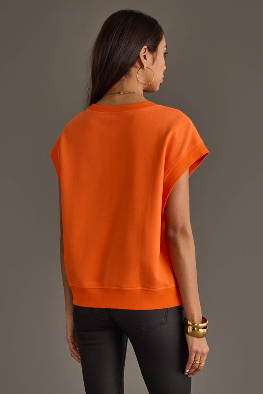 A woman stands facing away against a plain gray background, wearing the Panache Apparel Collection Orange & White Game Day Short Sleeve Sweatshirt, paired with black pants, gold hoop earrings, and gold bracelets.