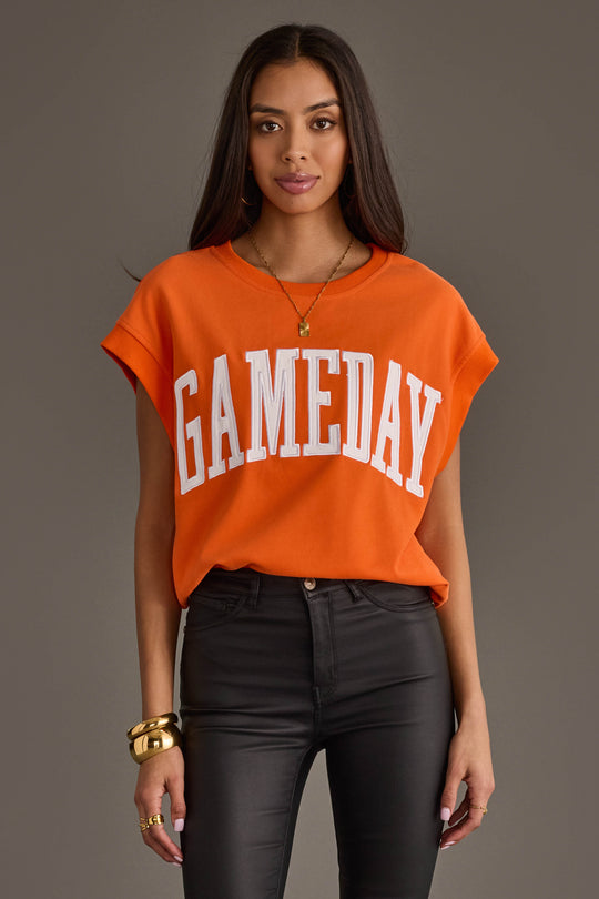 A woman wearing the Panache Apparel Collection Orange & White Game Day Short Sleeve Sweatshirt, featuring bold white "GAMEDAY" lettering, paired with black pants and gold jewelry, stands against a plain gray background.