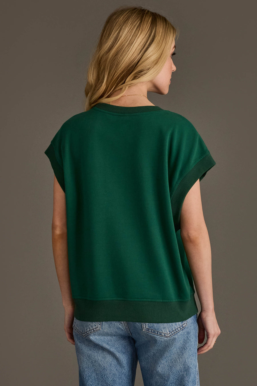 A person with long blonde hair, wearing the Panache Apparel Collection's Green & Gold Game Day Short Sleeve Sweatshirt and light blue jeans, is standing with their back to the camera against a plain gray background.