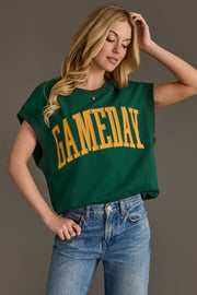 A woman stands against a plain background, posing with one hand on her hip and the other touching her hair, wearing the Panache Apparel Collection's Green & Gold Game Day Short Sleeve Sweatshirt paired with blue jeans.
