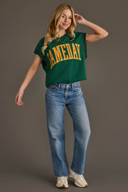 A woman stands smiling in a Green & Gold Game Day Short Sleeve Sweatshirt from Panache Apparel Collection, blue jeans, and white sneakers against a plain gray background.