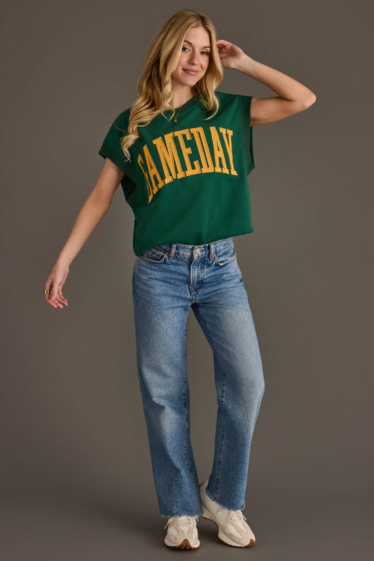 A woman stands smiling in a Green & Gold Game Day Short Sleeve Sweatshirt from Panache Apparel Collection, blue jeans, and white sneakers against a plain gray background.