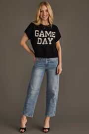 Smiling woman with blonde hair wearing the Panache Apparel Collection Black & White Game Day Short Sleeve Sweater, light blue jeans, and black open-toe heels, standing against a plain gray background.