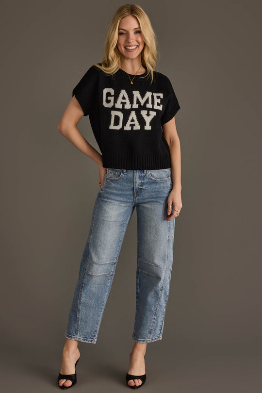 Smiling woman with blonde hair wearing the Panache Apparel Collection Black & White Game Day Short Sleeve Sweater, light blue jeans, and black open-toe heels, standing against a plain gray background.