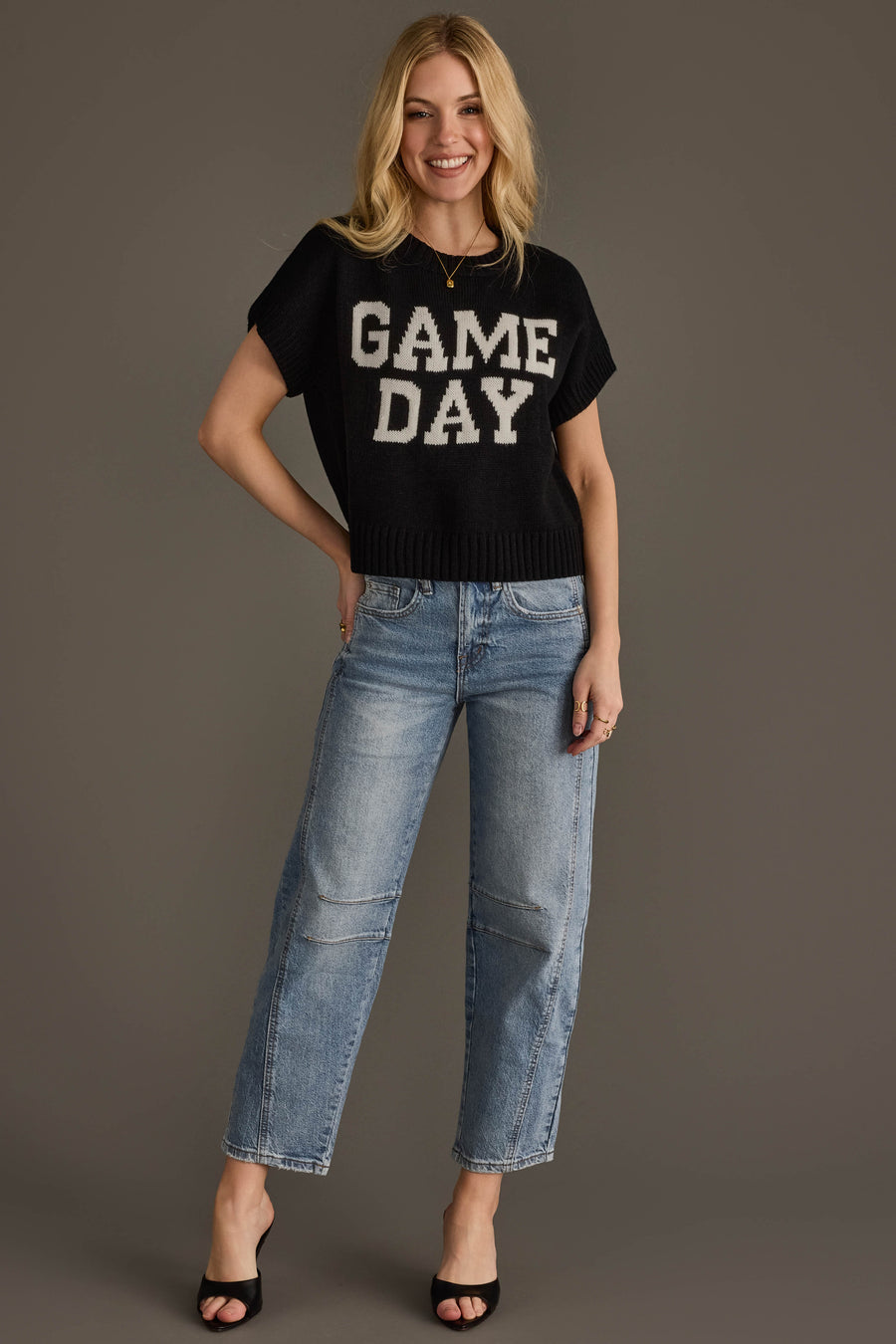 Smiling woman with blonde hair wearing the Panache Apparel Collection Black & White Game Day Short Sleeve Sweater, light blue jeans, and black open-toe heels, standing against a plain gray background.