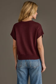 A woman with blonde hair, wearing the Panache Apparel Collection's Wine Season Short Sleeve Sweater and blue jeans, stands facing away from the camera against a plain gray background.