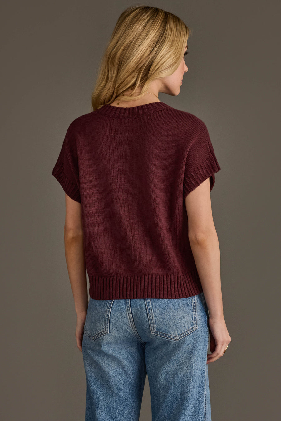 A woman with blonde hair, wearing the Panache Apparel Collection's Wine Season Short Sleeve Sweater and blue jeans, stands facing away from the camera against a plain gray background.