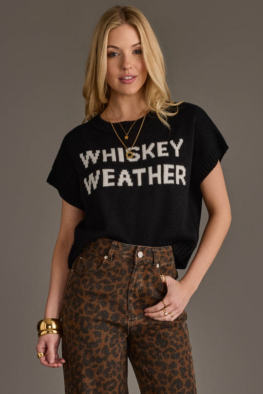 A woman with long blonde hair wears a Panache Apparel Collection Black & White Whiskey Weather Short Sleeve Sweater, leopard-print pants, gold bracelets, and layered necklaces as she stands against a plain gray background.