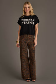 A woman with blonde hair stands against a gray background, smiling slightly. She is wearing the Panache Apparel Collection's Black & White Whiskey Weather Short Sleeve Sweater, paired with wide-leg brown leopard print pants, black peep-toe shoes, and gold bracelets.