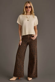 A woman stands against a gray background wearing sunglasses, an Ivory Short Sleeve Sweater from Panache Apparel Collection, a gold necklace, wide-leg brown leopard print pants, beige boots, and gold bracelets.