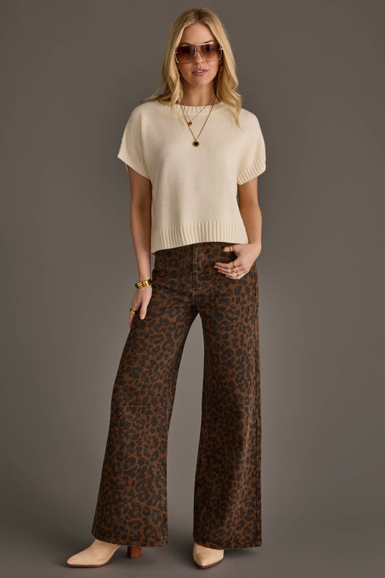 A woman stands against a gray background wearing sunglasses, an Ivory Short Sleeve Sweater from Panache Apparel Collection, a gold necklace, wide-leg brown leopard print pants, beige boots, and gold bracelets.