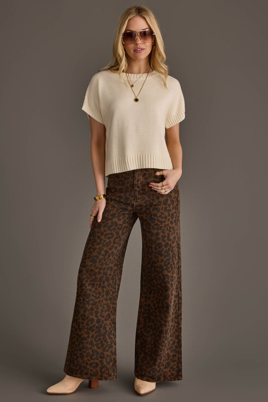 A woman stands against a gray background wearing sunglasses, an Ivory Short Sleeve Sweater from Panache Apparel Collection, a gold necklace, wide-leg brown leopard print pants, beige boots, and gold bracelets.