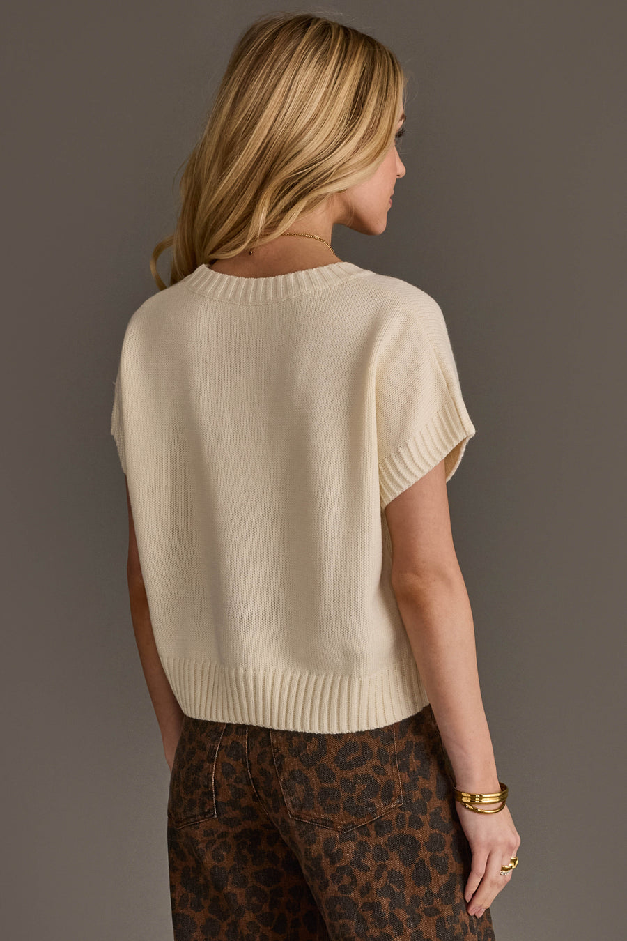 A woman with long blonde hair, seen from behind, wears the Panache Apparel Collection Ivory Short Sleeve Sweater paired with high-waisted brown leopard print pants. She is standing against a plain gray background.