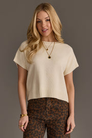 A woman with long blonde hair wears the Ivory Short Sleeve Sweater by Panache Apparel Collection, styled with layered necklaces, bracelets, and high-waisted leopard print pants. She stands against a plain gray background, smiling slightly.