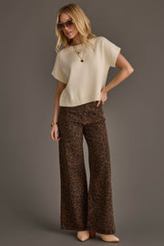 Dressed in the Panache Apparel Collection Ivory Short Sleeve Sweater, the woman poses against a plain gray background. She pairs the top with brown wide-leg leopard print pants, beige heels, and gold jewelry. Her blonde hair is styled in loose waves, and she completes the chic look with sunglasses.