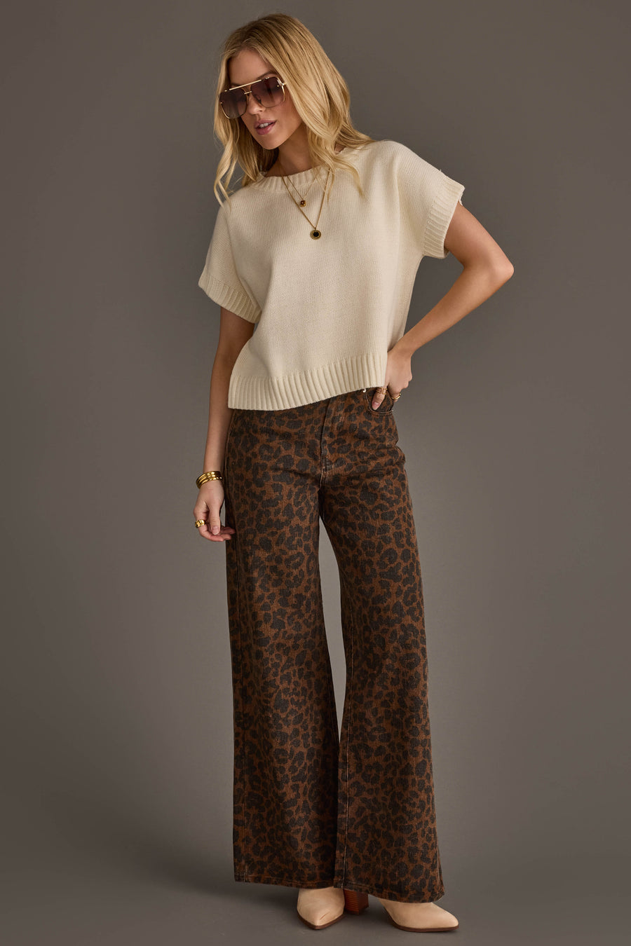 Dressed in the Panache Apparel Collection Ivory Short Sleeve Sweater, the woman poses against a plain gray background. She pairs the top with brown wide-leg leopard print pants, beige heels, and gold jewelry. Her blonde hair is styled in loose waves, and she completes the chic look with sunglasses.