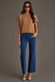 A woman stands against a plain background wearing sunglasses, the Panache Apparel Collection Light Brown Short Sleeve Sweater, blue high-waisted jeans, and nude heels, with one hand in her pocket and the other relaxed by her side.