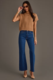 A woman stands against a plain gray background, wearing the Panache Apparel Collection Light Brown Short Sleeve Sweater, blue high-waisted jeans, beige heels, and sunglasses. She holds her sunglasses with one hand while looking toward the camera.