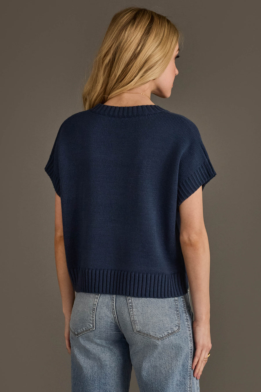 A woman with blonde hair is modeling the Panache Apparel Collection Navy Short Sleeve Sweater and light blue jeans, standing with her back to the camera against a plain brown background.