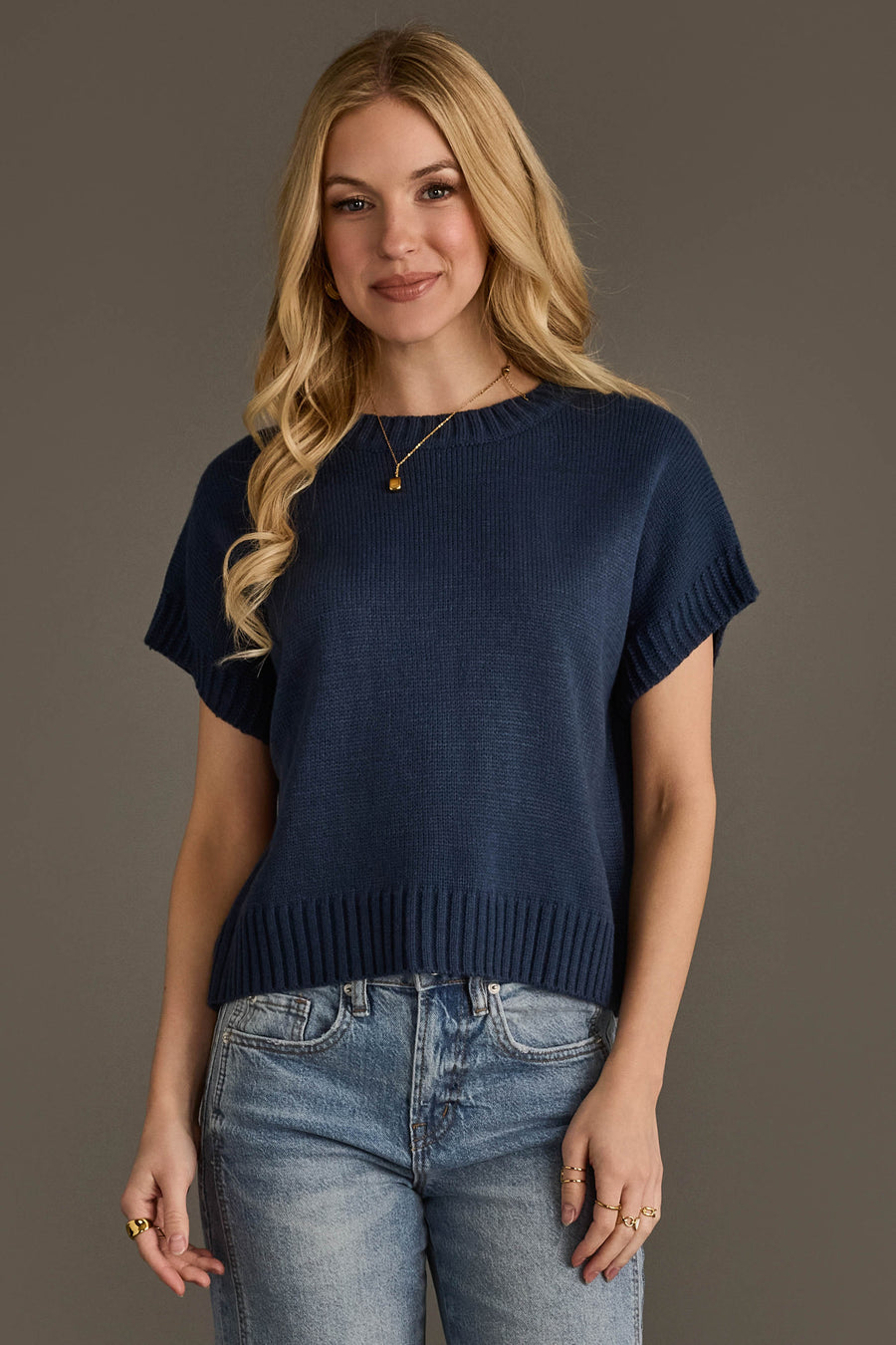 A person with long blonde hair is wearing the Panache Apparel Collection Navy Short Sleeve Sweater and light blue jeans, standing against a plain gray background and smiling gently at the camera.