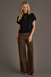 A woman with long blonde hair and a neutral expression stands against a gray background, wearing the Panache Apparel Collection Black Short Sleeve Sweater, paired with wide-leg brown leopard print pants, black heels, and accessorized with gold bracelets, necklaces, and rings.