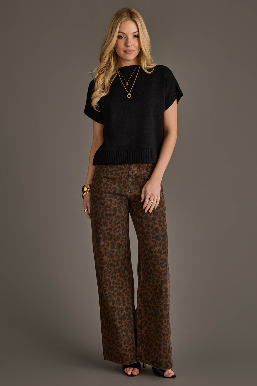 A woman with long blonde hair and a neutral expression stands against a gray background, wearing the Panache Apparel Collection Black Short Sleeve Sweater, paired with wide-leg brown leopard print pants, black heels, and accessorized with gold bracelets, necklaces, and rings.