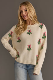 A woman wearing the Ivory Tree Sweater with Pink Bows, a cream-colored sweater featuring a repeating pattern of green sprigs tied with pink bows.