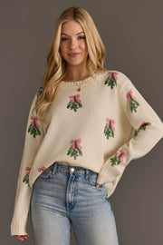 A person wears the Ivory Tree Sweater with Pink Bows, a cream-colored knit sweater patterned with repeating images of green mistletoe sprigs tied with pink bows.