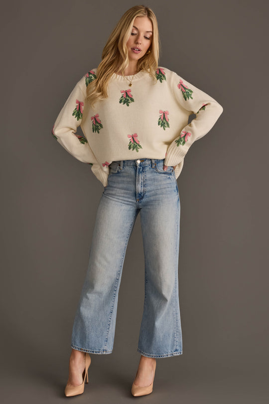 A person wearing an Ivory Tree Sweater with Pink Bows, a cream-colored knit with green mistletoe sprigs, paired with wide-leg light-wash jeans and nude pumps.