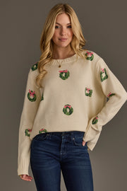 A woman wears an Ivory Wreath Sweater with Pink Bows from Panache Apparel Collection, an ivory crewneck sweater featuring a repeating pattern of green wreaths with pink bows.