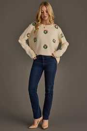 A woman wears an Ivory Wreath Sweater with Pink Bows in cream with small green wreaths and pink bows, paired with dark blue jeans and tan pumps.