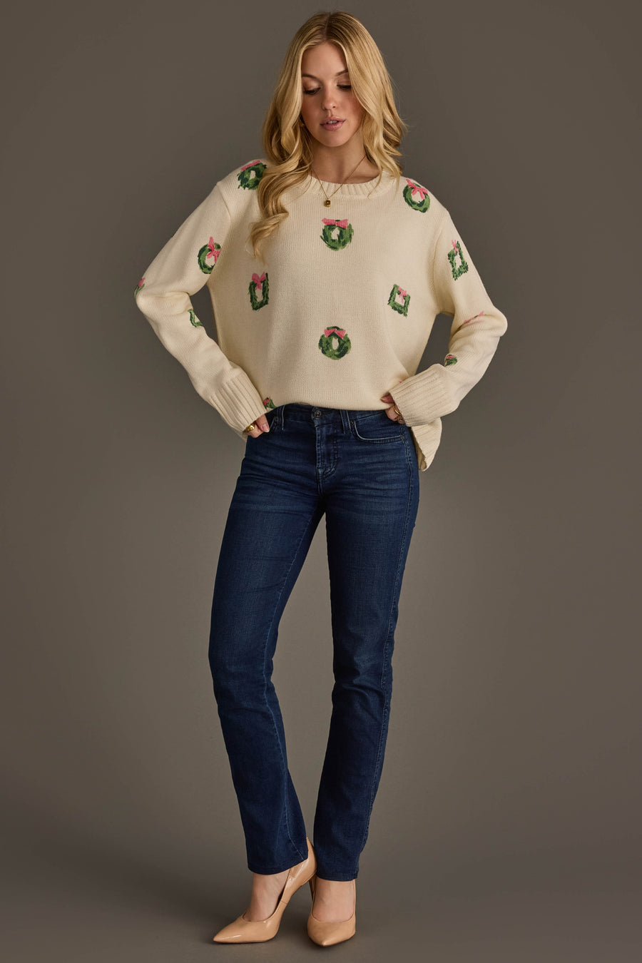 A woman wears an Ivory Wreath Sweater with Pink Bows in cream with small green wreaths and pink bows, paired with dark blue jeans and tan pumps.