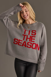 A woman smiles while wearing a gray knit Gray Tis The Season Sweater with TIS THE SEASON in red letters on the front.