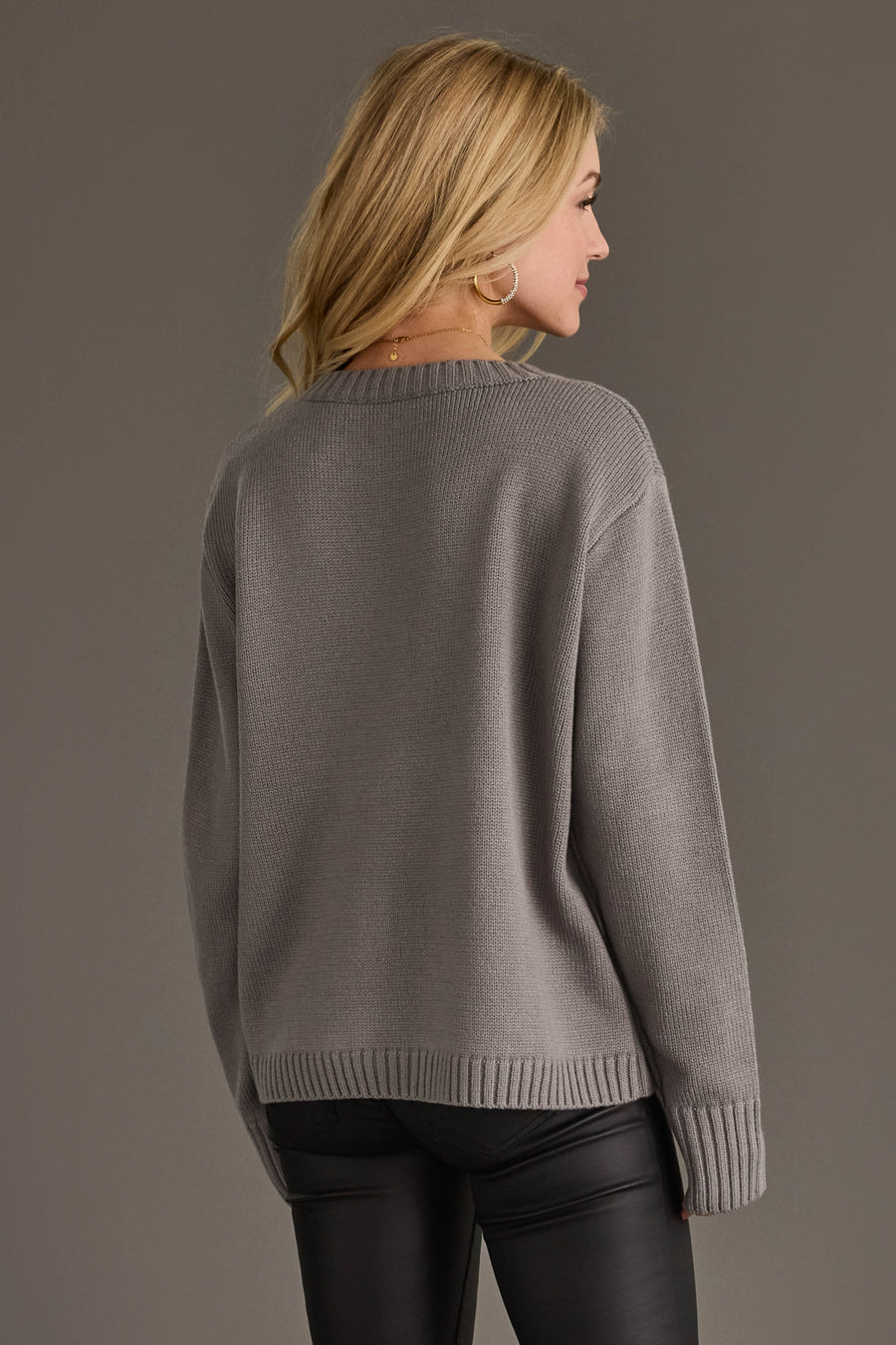 A person seen from the back wearing a long-sleeved, gray Gray Tis The Season Sweater and black pants.