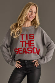 Woman wearing a Gray Tis The Season Sweater, featuring a gray crewneck with the text TIS THE SEASON in red letters across the front.