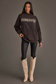 A woman with long dark hair stands in a relaxed pose against a gray background, wearing an oversized Panache Apparel Collection Dark Gray Day Drinking & Hungover Reversible Sweatshirt, black pants, and beige cowboy boots.
