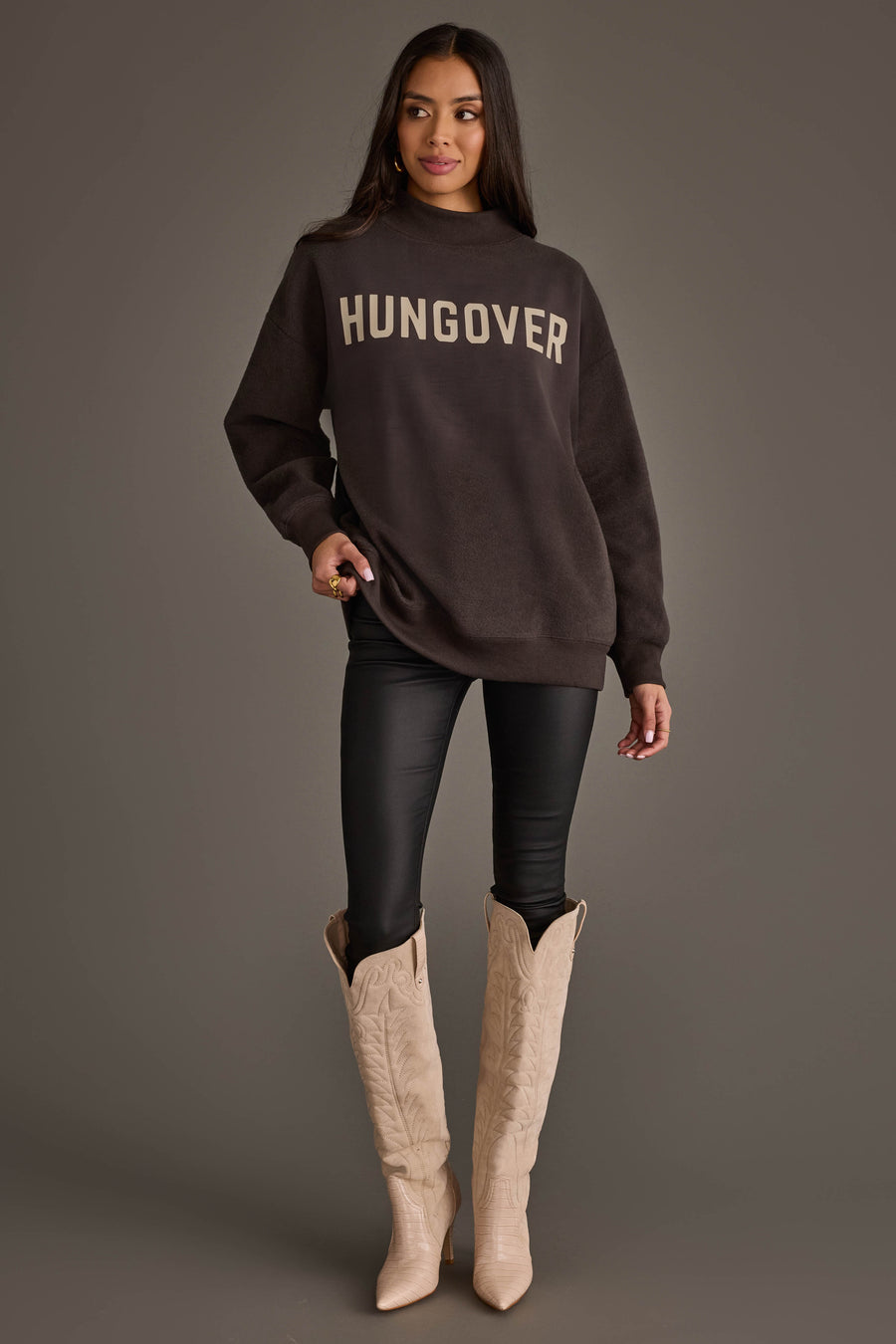 A woman with long dark hair stands in a relaxed pose against a gray background, wearing an oversized Panache Apparel Collection Dark Gray Day Drinking & Hungover Reversible Sweatshirt, black pants, and beige cowboy boots.