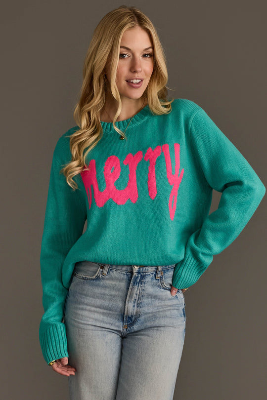 A person wears a light blue & bright pink merry sweater with the word merry written in large, bright pink script across the chest.