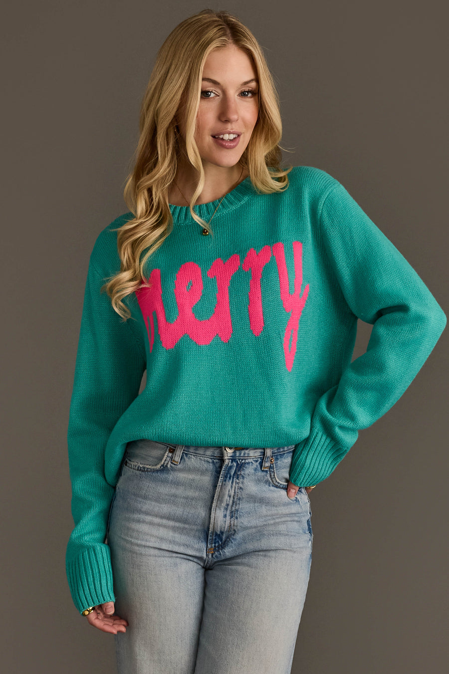 A person wears a light blue & bright pink merry sweater with the word merry written in large, bright pink script across the chest.