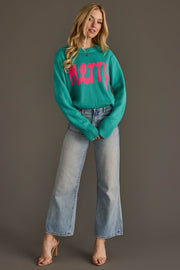 A woman wears the Light Blue & Bright Pink Merry Sweater, a teal long-sleeve top with pink lettering, paired with light-wash wide-leg jeans and heeled sandals.